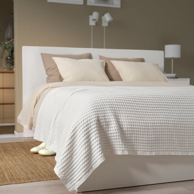 Neatly arranged bed with PELARTUJA cotton waffle bedspread, multiple pillows, headboard, lamp, slippers.