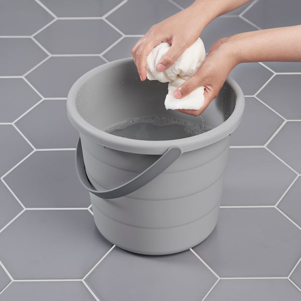 Person wrings out cloth over grey PEPPRIG bucket on tiled floor, showing its cleaning and water storage use.