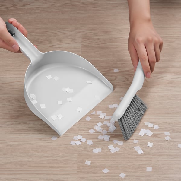 Person cleans floor with white PEPPRIG dustpan and brush, gathering small paper pieces; shows angle design effective for corner cleaning.
