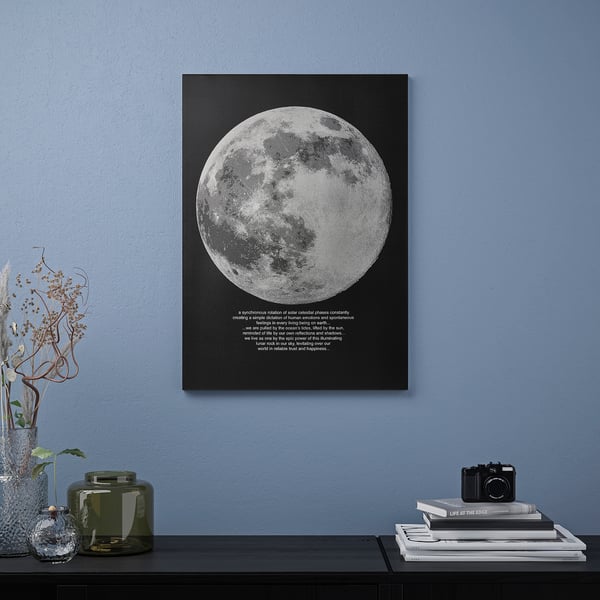 Black poster with large white moon hangs on blue wall next to shelf with books and vases.