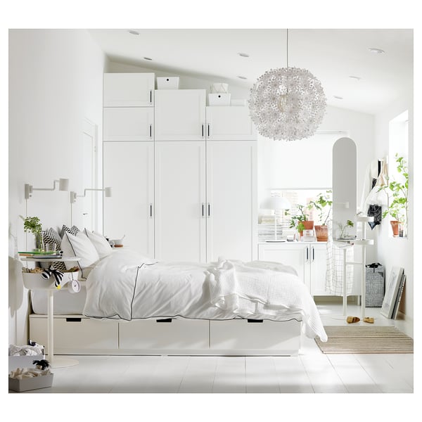Modern white bedroom with sleek PLATSA storage units, a bed with crisp white linens, stylish lighting, and plants.