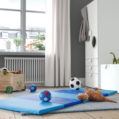 Kids playroom with blue PLUFSIG mats, toys, and storage. Soft, padded mats, white radiator, grey curtains, wooden floor.