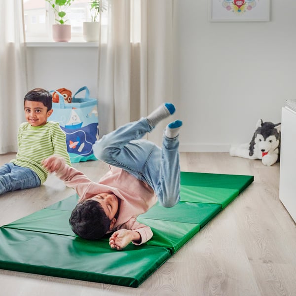 Two kids playing indoors, one doing cartwheels on a green foldable gym mat, enhancing motor skills and coordination.