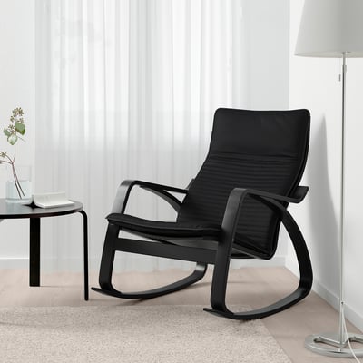 Black rocking chair on a rug, next to a small table with a vase and lamp.