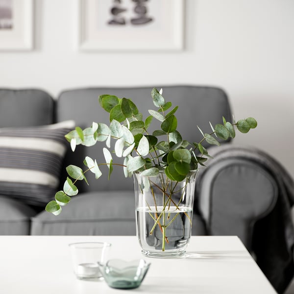 A glass POMP lantern vase with eucalyptus on a table, candles to the side, behind a grey sofa against a white wall.
