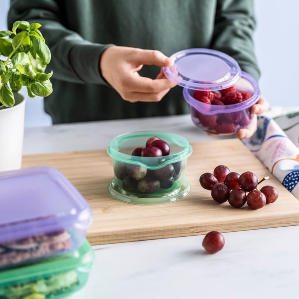 A person places the lid on a container holding grapes. On a wooden board, there are more grapes and silicone containers with lids for storing food.