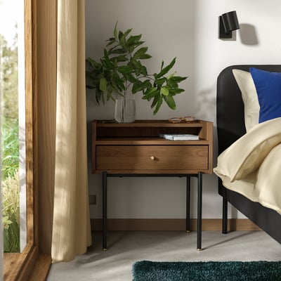 Bedroom scene featuring mid-century RÅDMANSÖ bedside table in warm walnut with black metal legs, cream curtains, and a blue pillow on a bed.