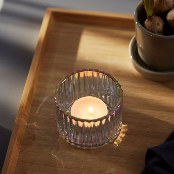 Small glass candle holder with ridged design sits on wooden table, reflecting light gently. Lit candle inside, casting soft shadows.