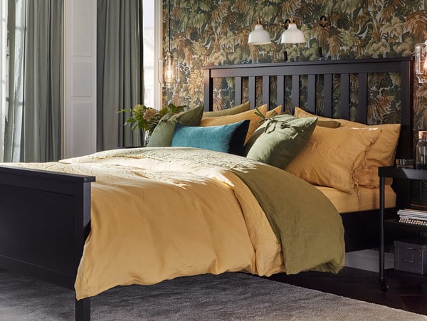 Bedroom with large bed, yellow and green bedding, dark wooden headboard, floral wallpaper, and adjustable RANARP lamps.