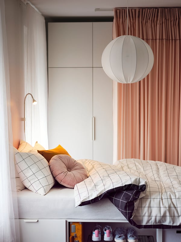 Cosy bedroom with light-colored bedding, colourful pillows, white pendant light, and yellow box beneath.