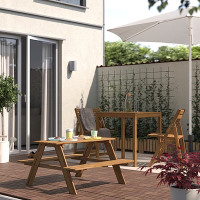 Small wooden RESÖ picnic table on deck, surrounded by matching chairs, near house with large window, perfect for kids.