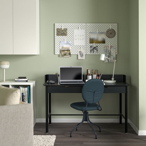 Modern home office setup: black rectangular desk, blue chair, green walls. Desk holds laptop, lamp, and supplies. Pegboard with decor above desk.