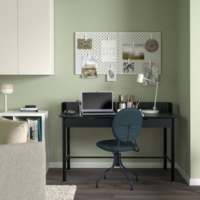 Modern home office setup: black rectangular desk, blue chair, green walls. Desk holds laptop, lamp, and supplies. Pegboard with decor above desk.