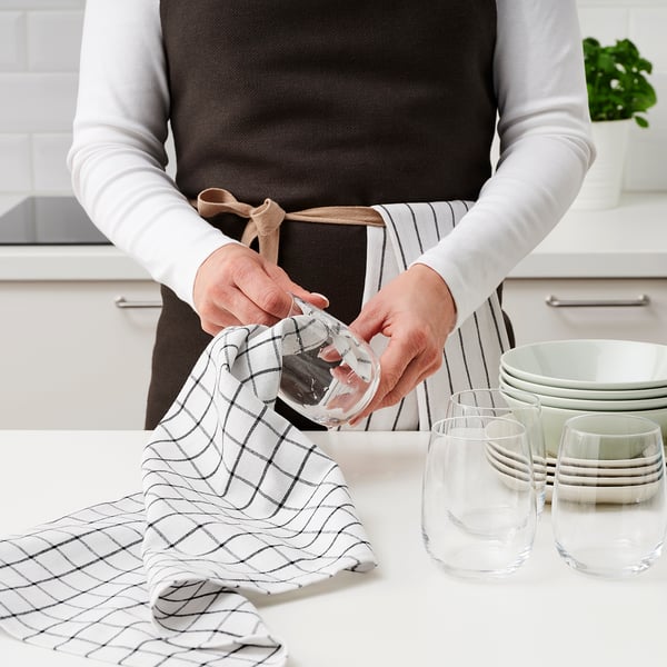 Person drying glass with a chequered towel on a kitchen counter near wine glasses and plates.