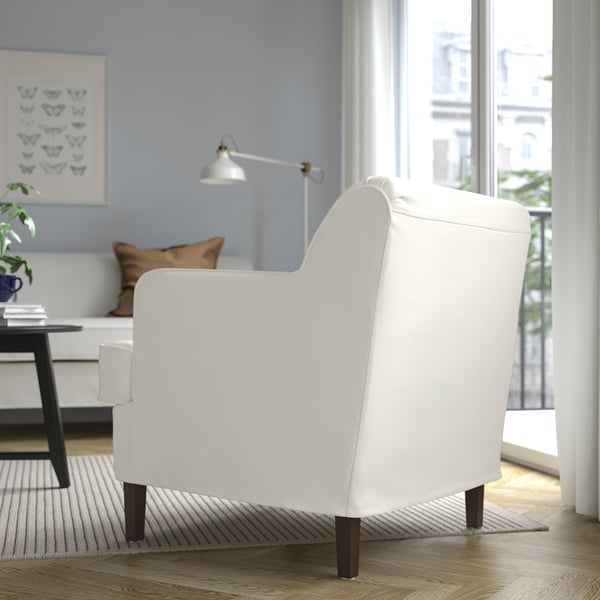 A bright, modern living room with a white ROCKSJÖN armchair, featuring wooden legs and a backrest angled for comfort. Large windows offer a city view.