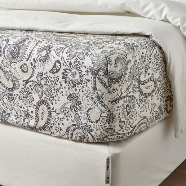 A bed with a cream and grey paisley printed fitted sheet, neatly fitted, displaying its elastic edging. White pillows rest on it.