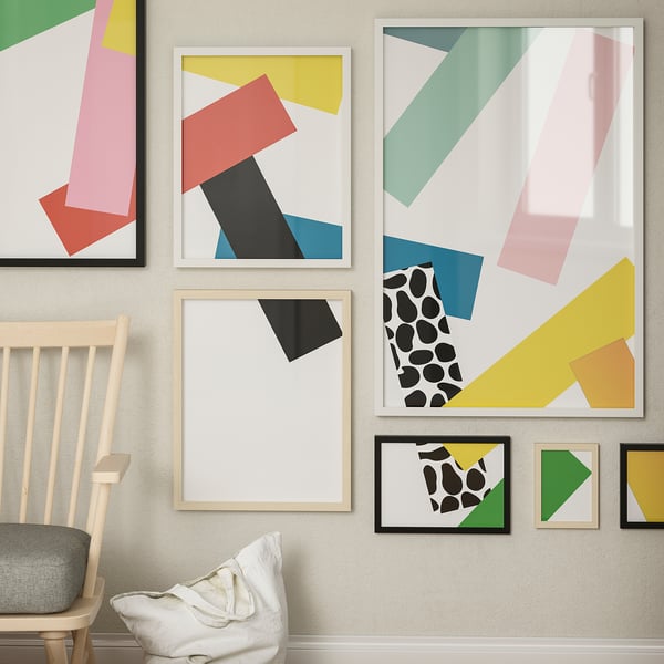 Modern art gallery wall with colorful geometric prints in mixed frames, wooden chair with cushion.