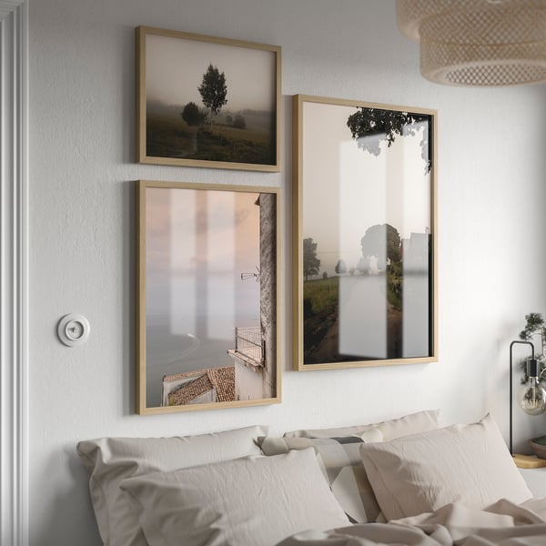 Three light wooden frames above bed show landscapes. Largest on right, smaller ones left. White & grey bedding, wicker pendant lamp above.