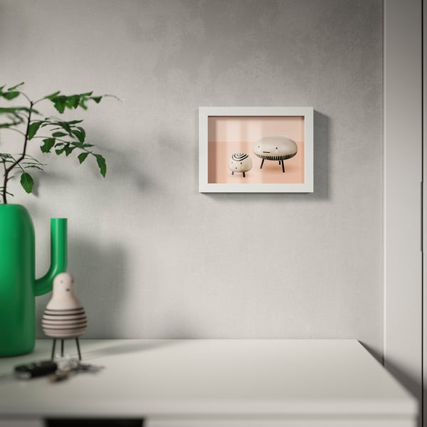 A modern white picture frame hangs on a grey wall, displaying a pink-toned minimalist furniture. Below it, a desk holds a green vase with branches.