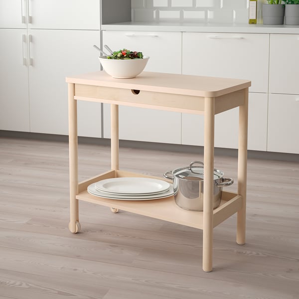 RÖNNINGE light wooden kitchen trolley with bowl, plates, and pots. Compact, two-tier design for extra storage. Fits well beside white cabinets.