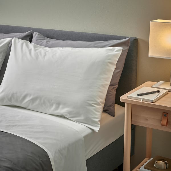 White bedding set with pillowcases on bed, grey headboard, wooden bedside table with notepad, lamp.