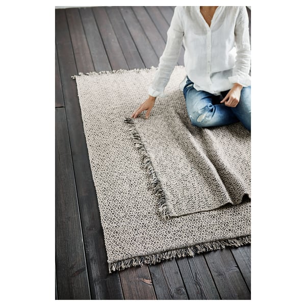 Person arranging a beige and black RÖRKÄR cotton rug on dark wooden floor.