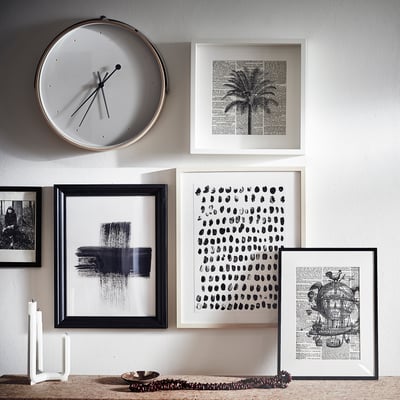 A white silent clock with black hands hangs above a wall of various framed black and white artworks.