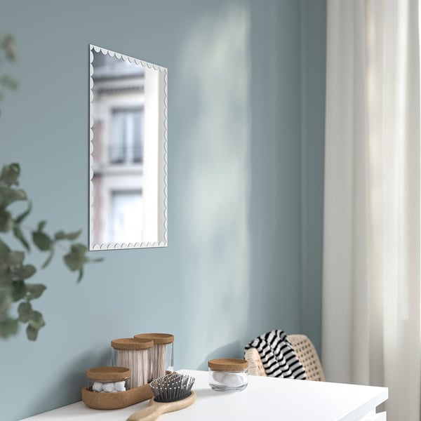 Clear rectangular mirror with wavy edges on light blue wall. On dresser, a brush, jars, and plant. Easy to hang with tape.