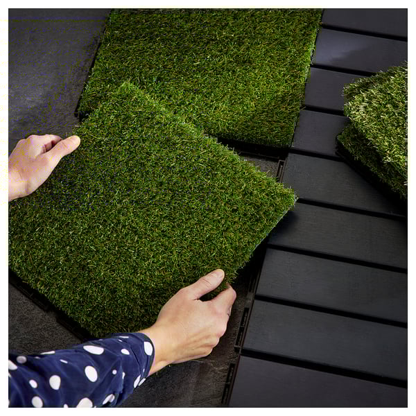 Person placing green artificial grass panels on dark slatted decking floor.