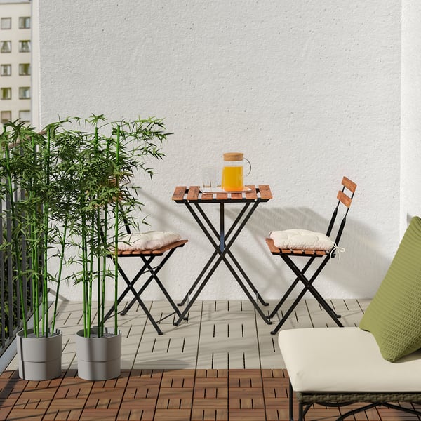 Cosy balcony with RUNNEN decking, small wooden table and chairs, lush bamboo plant, and white wall backdrop.