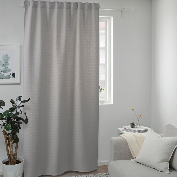 Long light gray RYSSBRÄKEN curtain hangs near white couch, blocking drafts. Patterned, with a rod at top, its taller than nearby plants.
