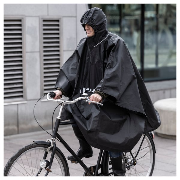 Person wearing black SALTSTEN rain poncho rides a bicycle in the city, designed to keep the person dry with a hood and front pocket.