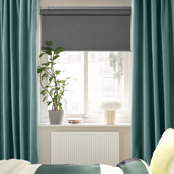 Large window with grey SANELA curtains, dense fabric, darkening light, plant on ledge, lamp, bed below.