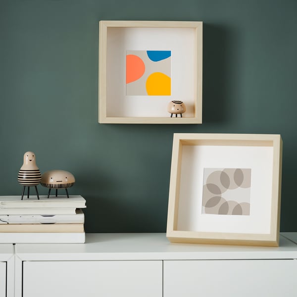 Two framed artworks hang on green wall: one colourful, one geometric. Shelves below hold books and sculptures, creating shadows.