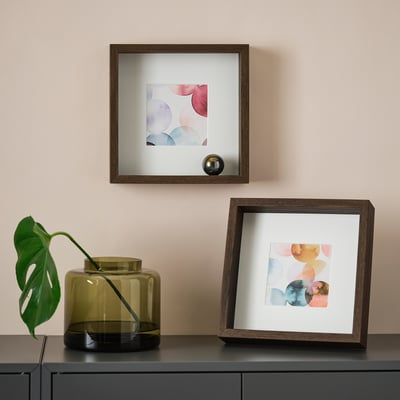 Two abstract art frames on wall, one held by stand. Plant in glass vase below.