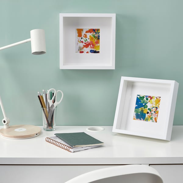 A well-lit desk with colourful framed art, a lamp, stationery, and notepads, showcasing a useful and aesthetically pleasing workspace setup.