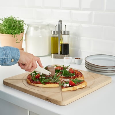 Person slicing pizza on rustic wooden board, which has a groove to catch juices. On a clean kitchen counter.