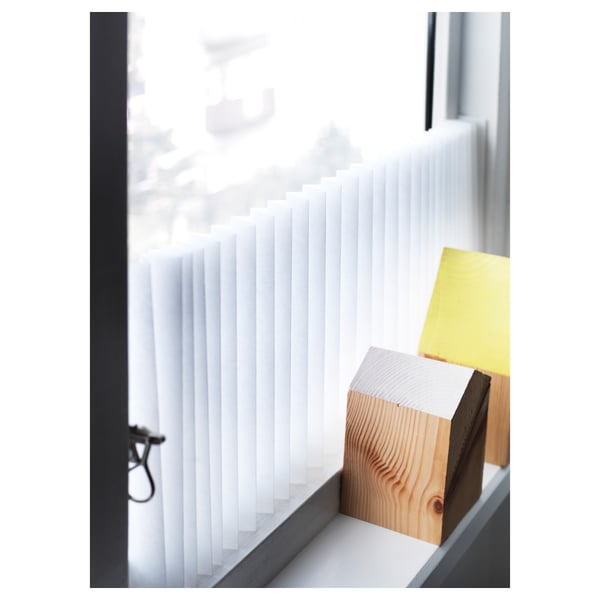 White SCHOTTIS blind, partially drawn, with wooden block and yellow paper support, mounted on window frame for privacy and light.