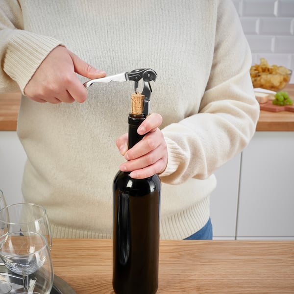 Person opens wine bottle with corkscrew. Scene on wooden table with glasses.