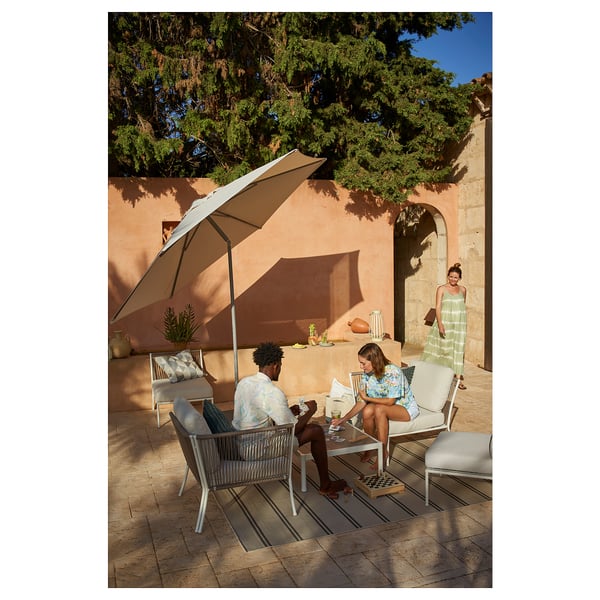 Two people sit under a large beige patio umbrella on modern white wicker outdoor furniture, enjoying a sunny day.