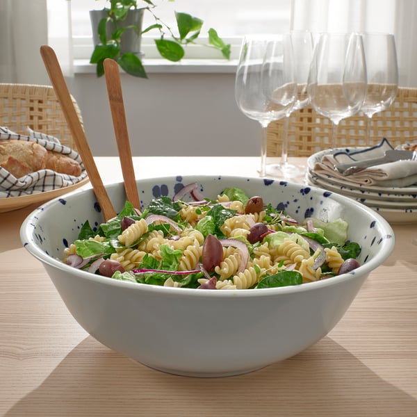Wooden salad tongs rest in a bowl of pasta salad, surrounded by plates and wine glasses.