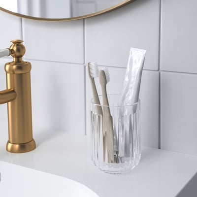 Clear glass toothbrush holder, holding toothbrushes and toothpaste, sits near gold tap.
