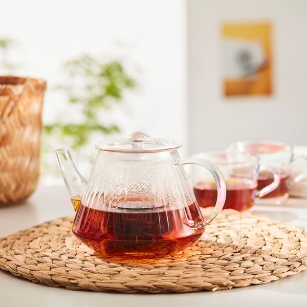 Clear glass teapot with infuser sits on woven mat, filled with red tea, two cups beside it.