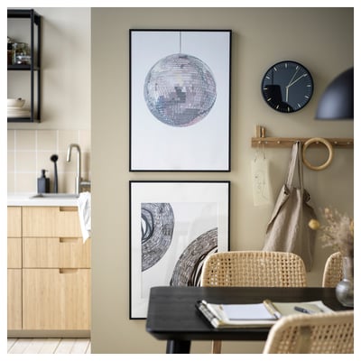 Cosy room with wooden furniture, a black clock, and modern art frames. A woven chair and a small desk with a notebook sit under a wall decoration.