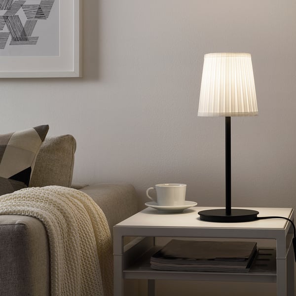 Small SKAFTET black lamp on table next to coffee cup, beside beige couch.