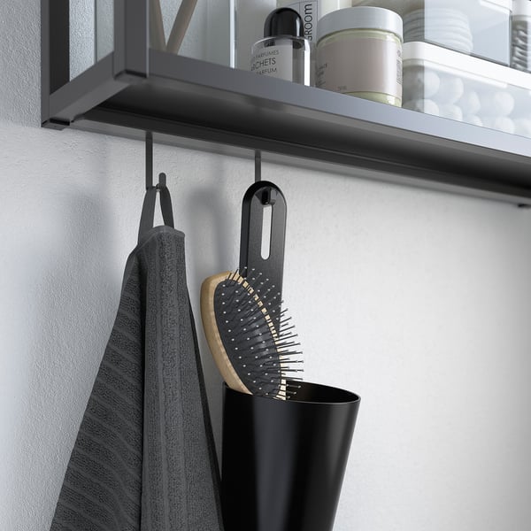 Black SKATTÅN container hangs on wall hook, holding items like a hairbrush and grey towel. Suitable for bathroom or kitchen storage.