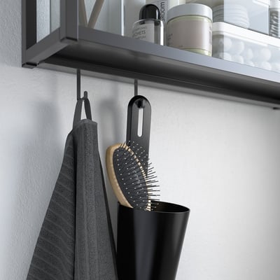 Black SKATTÅN container hangs on wall hook, holding items like a hairbrush and grey towel. Suitable for bathroom or kitchen storage.
