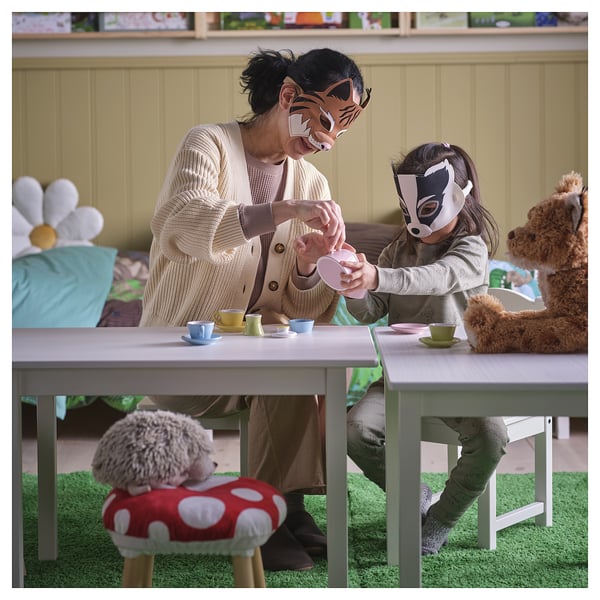 An an adult and child playing with SKOGSDUVA masks, sitting at a small table with tea cups, surrounded by stuffed animal toys.