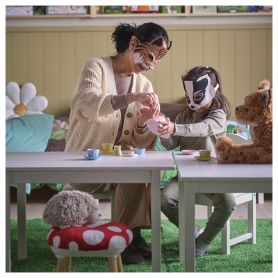 An an adult and child playing with SKOGSDUVA masks, sitting at a small table with tea cups, surrounded by stuffed animal toys.