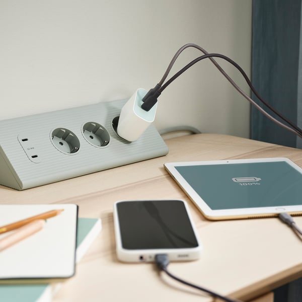 Wooden desk with white SKOTAT extension cord, black charging cable, smartphone, ipad, notebook, pencils.